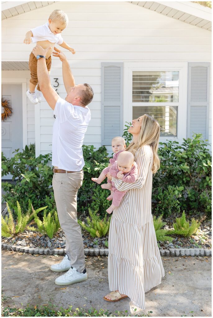 Family of five outside their home in Tampa, Florida. Mom holds twin baby girls while dad throws toddler boy up in the air